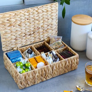 Puede incluir: Una caja de almacenamiento rectangular de pasto marino trenzado con tapa abatible, que cuenta con ocho compartimentos internos llenos de varias bolsitas de té, pequeños dulces envueltos y paquetes individuales. El material tiene una textura trenzada.