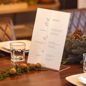May include: A clear acrylic menu holder displaying a menu with German text. The menu is divided into sections: Vorspeise, Hauptgericht, and Dessert. The holder is on a wooden table with decorative elements.