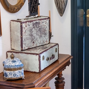 Pode incluir: Duas malas retangulares com uma estampa de mapa-múndi vintage em tons de creme e castanho escuro, com fechos e pegas de metal. Ao lado, encontra-se uma pequena e ornamentada caixa de porcelana azul e branca com acabamentos em metal dourado.
