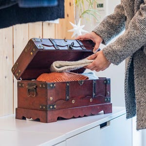 Pode incluir: Um baú de madeira vintage com tampa articulada, detalhes em latão e mancha escura. O baú está aberto, revelando roupas dobradas no interior. O baú está sobre uma superfície branca.