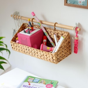 Op de afbeelding: Een rechthoekige hangmand van gevlochten zeegras, opgehangen aan een lichthouten wandstang. De mand bevat een roze kubusvormige luidspreker, een lang potlood met een regenboogontwerp, een rode stift en witte pennen. Witte haken hangen aan de stang.