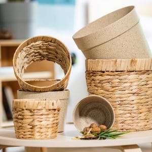 Op de afbeelding: Een verzameling decoratieve plantenbakken en manden. De afbeelding toont verschillende beige gespikkelde keramische potten en geweven manden van natuurlijke vezels. De potten zijn gestapeld en sommige zijn leeg, andere bevatten kleine items.