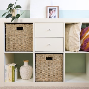 May include: A white cube storage unit with two woven baskets, two white drawers, and decorative pillows. A ceramic vase and books are on a shelf. A framed photo and a potted plant are on top.