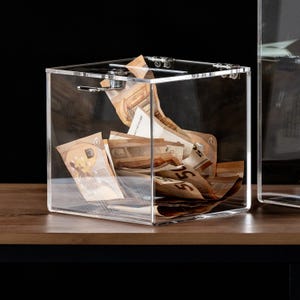 May include: A clear acrylic donation box filled with Euro banknotes. The box is square with a hinged lid and metal hardware. The banknotes are visible through the transparent sides, creating a visual display of the contents.