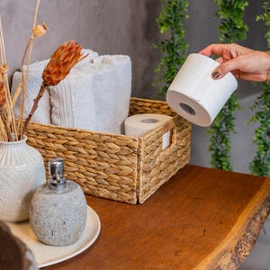 Pode incluir: Uma cesta retangular de vime trançado contendo toalhas brancas dobradas e um rolo de papel higiênico, acompanhada por um vaso de cerâmica branca com flores secas laranja e um dispensador de sabonete de pedra cinza.