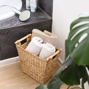 Op de afbeelding: Een rechthoekige mand van gevlochten zeegras met twee lichtgekleurde houten handvatten. De mand bevat drie opgevouwen beige en witte katoenen handdoeken. De mand heeft een gestructureerd vlechtpatroon en staat op een lichte houten vloer.
