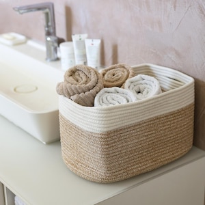 Peut inclure: Un panier de rangement rectangulaire en corde tressée avec un design bicolore composé d'une base beige naturel et d'une partie supérieure crème. Le panier contient plusieurs serviettes roulées dans des tons beige, ocre et blanc cassé.