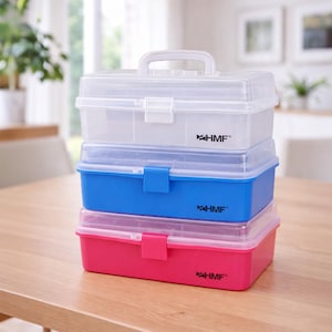 Può includere: Una pila di tre scatole di plastica per la conservazione in bianco, blu e rosa. Ogni scatola ha un coperchio trasparente e una maniglia sulla parte superiore. Le scatole sono impilate su un tavolo di legno. Il marchio "HMF" è stampato su ogni scatola.