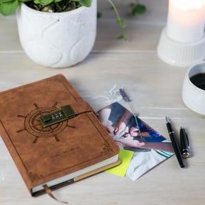 Può includere: Un diario in pelle marrone con lucchetto a combinazione, un design a ruota nautica e le parole "Premium Quality Notebooks Vintage Style". Sono visibili anche una foto, penne, una tazza di caffè, una candela e una pianta.
