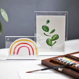 Könnte beinhalten: Zwei transparente Acryl-Fotorahmen. Einer zeigt einen Aquarell-Regenbogen in Rot, Orange und Gelb. Der andere zeigt ein Aquarellgemälde mit grünen Blättern. Ein hölzernes Aquarellfarbenset und ein Pinsel sind ebenfalls zu sehen.