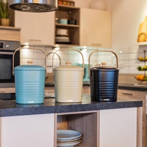 Könnte beinhalten: Drei Küchen-Komposteimer in Türkis, Creme und Schwarz, jeweils mit Holzgriff und Deckel. Die Eimer haben ein geripptes Design und ein kleines Logo auf der Vorderseite. Sie stehen auf einer Arbeitsplatte.