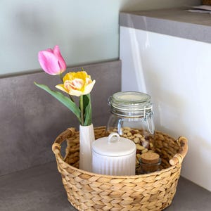 Op de afbeelding: Een ronde gevlochten mand van waterhyacint met een witte geribbelde keramische vaas met één roze en één gele tulp, een witte geribbelde keramische pot met deksel, een glazen pot met metalen beugelsluiting gevuld met noten en een klein glazen potje met kurk.