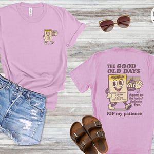 Puede incluir: Una camiseta rosa claro con un gráfico en la parte delantera y trasera. El gráfico frontal es un personaje de boleto de dibujos animados. El gráfico trasero dice "THE GOOD OLD DAYS MOUNTAIN" con un personaje de dibujos animados y texto. Pantalones cortos de mezclilla y sandalias también están en la imagen.