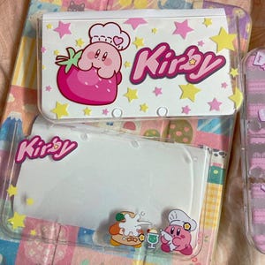 Könnte beinhalten: Transparente Nintendo 3DS-Hüllen aus Kunststoff mit farbenfrohen Kirby-Designs. Eine Hülle zeigt einen Erdbeer-Charakter und das Wort "Kirby" in Pink. Die andere Hülle hat einen weißen Hintergrund mit dem Wort "Kirby" in Pink.
