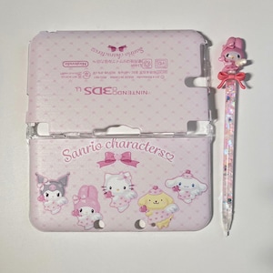 Op de afbeelding: Een roze Nintendo 3DS LL consolehoes en een bijpassende pen. De hoes heeft Sanrio-personages en de tekst "Sanrio characters". De pen heeft een roze karaktertop en een transparante schacht gevuld met glitter.