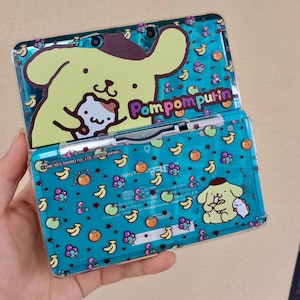 Könnte beinhalten: Eine türkisfarbene Nintendo 3DS-Handheld-Spielkonsole mit einer transparenten Schutzhülle mit einem gelben Cartoon-Hund, Früchten und dem Text "Pompompurin". Die Hülle hat ein sich wiederholendes Muster aus Bananen, Äpfeln und Orangen.