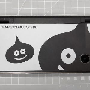May include: A black and white Nintendo DS Lite featuring a Dragon Quest IX design. The design includes the text "DRAGON QUEST IX" and two cartoon slime characters. The handheld console is on a gray surface.