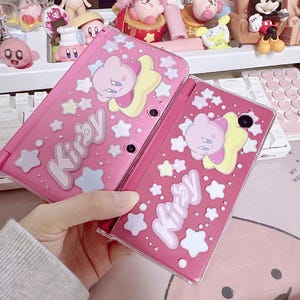 Puede incluir: Dos consolas Nintendo 3DS rosas con temática de Kirby. Las consolas presentan la palabra "Kirby" en escritura blanca, junto con gráficos de estrellas y personajes. Las consolas se sostienen en una mano.