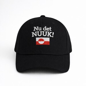 Puede incluir: Gorra de b&eacute;isbol negra con la inscripci&oacute;n blanca bordada "Nu det NUUK!". Debajo, una peque&ntilde;a bandera groenlandesa bordada, con un c&iacute;rculo blanco sobre fondo rojo.