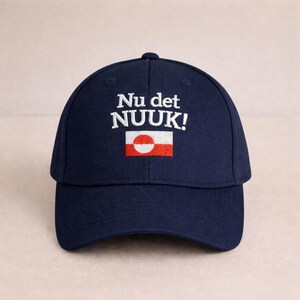 Puede incluir: Gorra de b&eacute;isbol azul marino con texto bordado en blanco "Nu det NUUK!" y un peque&ntilde;o emblema de bandera. La bandera presenta un c&iacute;rculo blanco y una franja roja. Un accesorio de moda para el uso diario.
