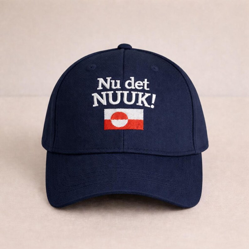 Puede incluir: Gorra de b&eacute;isbol azul marino con texto bordado en blanco "Nu det NUUK!" y un peque&ntilde;o emblema de bandera. La bandera presenta un c&iacute;rculo blanco y una franja roja. Un accesorio de moda para el uso diario.