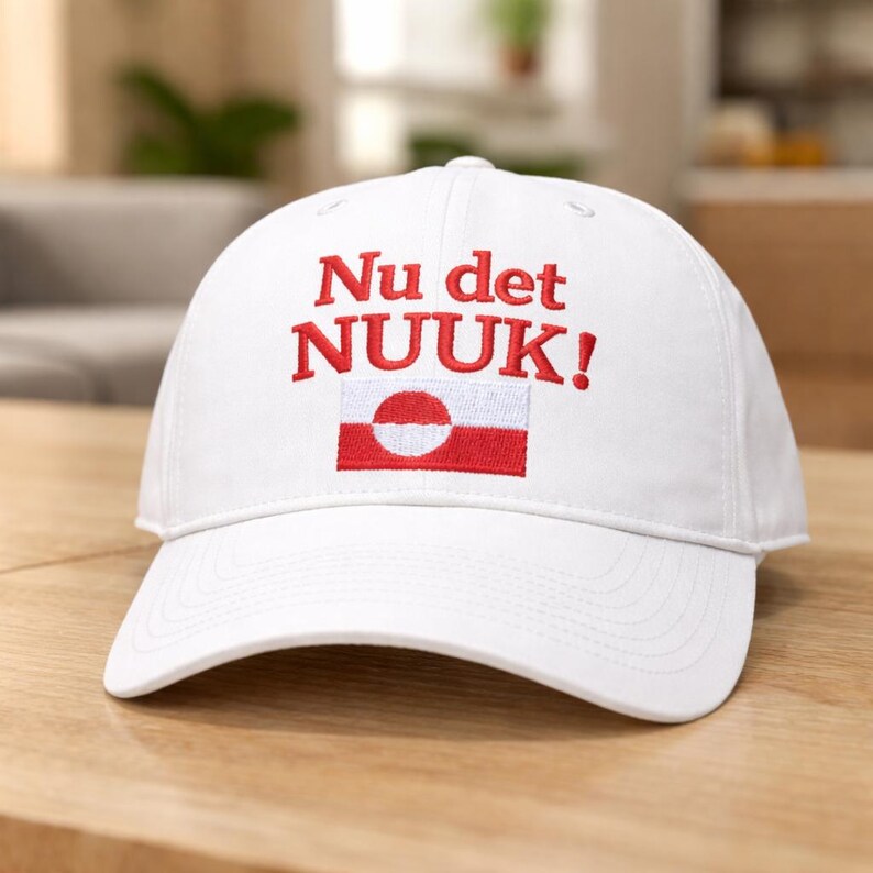 Puede incluir: Gorra de b&eacute;isbol blanca con el texto "Nu det NUUK!" bordado en rojo. La gorra presenta un emblema de bandera roja y blanca. El sombrero est&aacute; hecho de un material suave y tiene una visera curva.