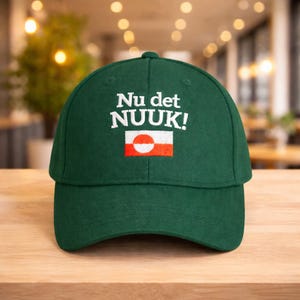 Puede incluir: Gorra de b&eacute;isbol verde oscuro con la inscripci&oacute;n "Nu det NUOK!" bordada en blanco. Debajo, un peque&ntilde;o dise&ntilde;o de bandera con un c&iacute;rculo blanco y un rect&aacute;ngulo rojo. La gorra est&aacute; sobre una superficie de madera.