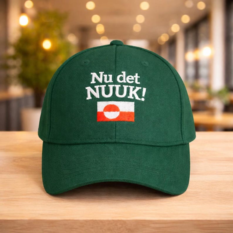 Puede incluir: Gorra de b&eacute;isbol verde oscuro con la inscripci&oacute;n "Nu det NUOK!" bordada en blanco. Debajo, un peque&ntilde;o dise&ntilde;o de bandera con un c&iacute;rculo blanco y un rect&aacute;ngulo rojo. La gorra est&aacute; sobre una superficie de madera.