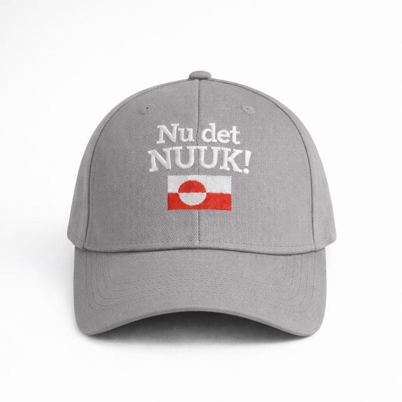 Puede incluir: Gorra de b&eacute;isbol gris con el texto "Nu det NUUK!" bordado en blanco sobre un peque&ntilde;o dise&ntilde;o de bandera. La bandera presenta un rect&aacute;ngulo rojo con un c&iacute;rculo blanco y un punto rojo.