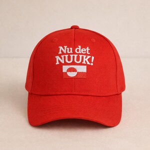 Puede incluir: Gorra de b&eacute;isbol roja brillante con el texto bordado "Nu det NUUK!" en blanco. Debajo del texto hay un peque&ntilde;o parche rectangular con un dise&ntilde;o blanco y rojo. La gorra tiene una visera curva y una corona redondeada.