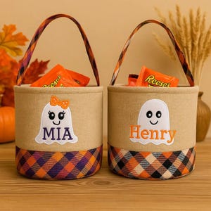 Op de afbeelding: Twee beige Halloween-traktatie-emmers met geruite bodems en handvatten. Eén emmer heeft een spook met een strik en de naam "MIA" erop geborduurd. De andere emmer heeft een spook en de naam "Henry". Beide emmers zijn gevuld met snoep.