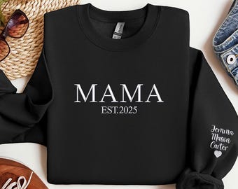 Könnte beinhalten: Schwarzes Sweatshirt mit dem weißen Wort "MAMA" und "EST.2025" darunter. Der Ärmel ist mit dem Namen "Jemima Mason Carter" bestickt. Eine Sonnenbrille und ein brauner Schuh sind im Hintergrund zu sehen.