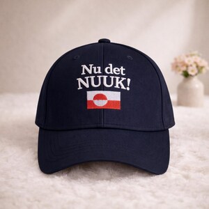 Puede incluir: Gorra de b&eacute;isbol azul marino con el texto "Nu det NUUK!" en blanco, sobre un peque&ntilde;o dise&ntilde;o de bandera. La bandera presenta un c&iacute;rculo blanco sobre fondo rojo, con una franja horizontal blanca. Un accesorio elegante.