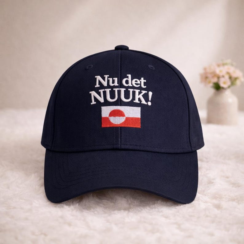 Puede incluir: Gorra de b&eacute;isbol azul marino con el texto "Nu det NUUK!" en blanco, sobre un peque&ntilde;o dise&ntilde;o de bandera. La bandera presenta un c&iacute;rculo blanco sobre fondo rojo, con una franja horizontal blanca. Un accesorio elegante.