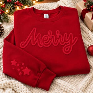 May include: A red crewneck sweatshirt featuring the word Merry in a raised, tonal script font across the chest. The left sleeve is decorated with a cluster of raised, tonal star shapes of varying sizes. The garment has a ribbed collar and cuffs.