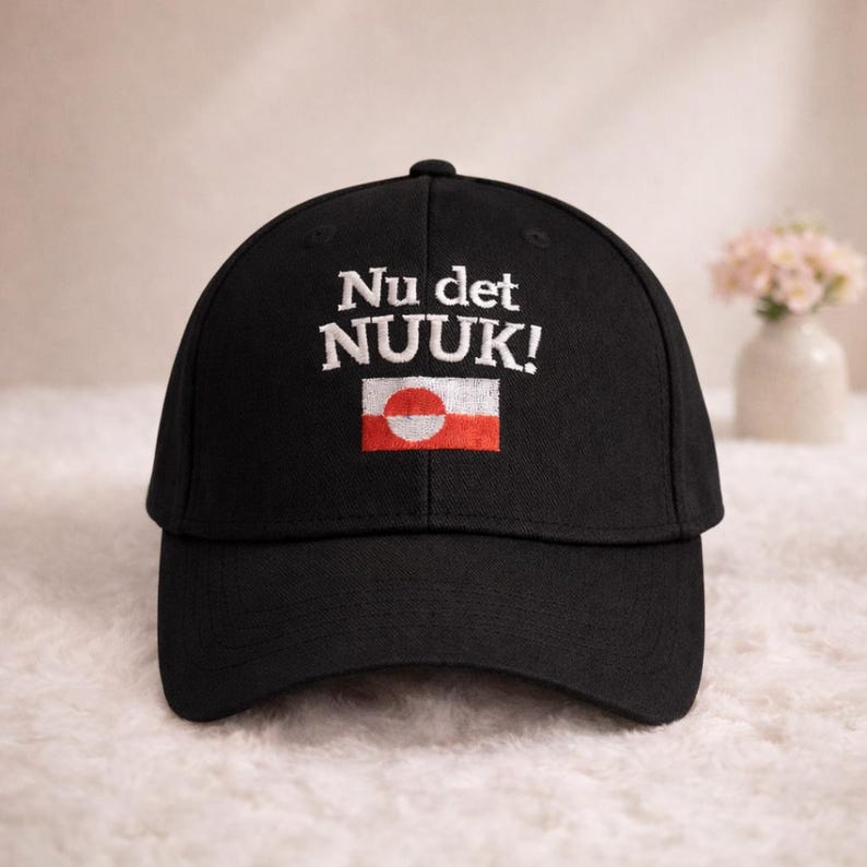 Puede incluir: Gorra de b&eacute;isbol negra con el texto "Nu det NUUK!" bordado en blanco. Debajo del texto, un peque&ntilde;o dise&ntilde;o rectangular con un c&iacute;rculo blanco sobre fondo rojo. La gorra tiene una visera curva.