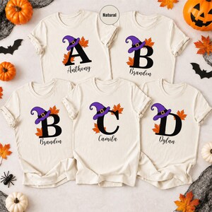 Könnte beinhalten: Fünf naturfarbene T-Shirts mit den schwarzen Buchstaben A, B, C und D. Jeder Buchstabe ist mit einem lila Hexenhut und orangefarbenen Ahornblättern verziert. Die Namen Anthony, Brandon, Camila und Dylan sind in schwarzer Schrift aufgedruckt.