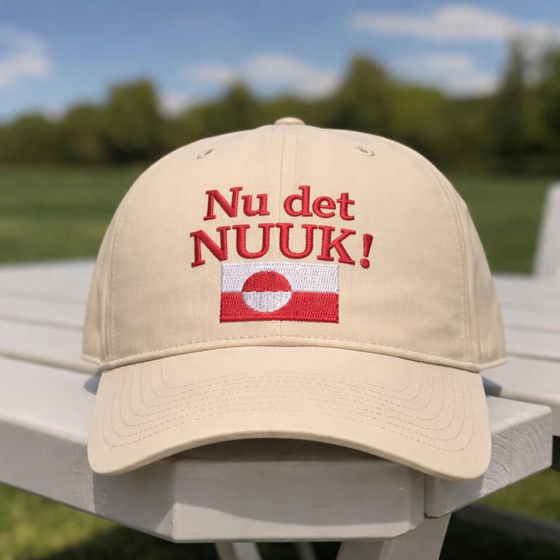 Puede incluir: Gorra de b&eacute;isbol beige con el texto bordado "Nu det NUUK!" en rojo, encima de un peque&ntilde;o dise&ntilde;o de bandera. La bandera presenta un c&iacute;rculo blanco sobre un fondo rojo, con una franja blanca en la parte superior e inferior.