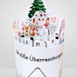 Może przedstawiać: Biały kalendarz adwentowy z 24 ponumerowanymi przegr&oacute;dkami. Zawiera ilustracje choinki, Świętego Mikołaja, renifera i innych świątecznych obraz&oacute;w. Na boku widnieje napis "24 s&uuml;&szlig;e &Uuml;berraschungen".