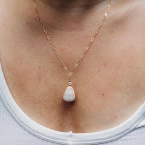 Puede incluir: Un delicado collar de cadena dorada con un colgante con una piedra blanca moteada en forma de lágrima y una pequeña perla. El collar se lleva alrededor del cuello, mostrando el diseño y el color de la joya.