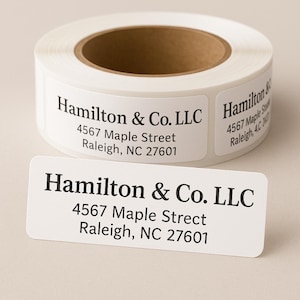 May include: A roll of white address labels with the text "Hamilton & Co. LLC" and address details in black. A single label is shown in front of the roll, also displaying the same information.