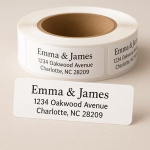 May include: A roll of white address labels with the text "Emma & James" and address details printed in black. A single label is shown below the roll, also displaying the same information.