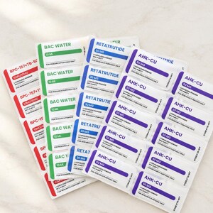 May include: A collection of rectangular labels arranged in a fan shape. The labels feature color-coded sections in red, green, blue, and purple, with printed text including BPC-157+TB-500, BAC WATER, RETATRUTIDE, and AHK-CU, along with dosage and purity details.