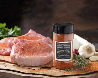 May include: A glass jar of blackened seasoning with a black lid. The label reads "Razy's Design & Creations Dry Rubs & Seasonings Blackened Seasoning Handcrafted | No Fillers Ingredients: Paprika, salt, black pepper, cayenne pepper, onion powder, garlic powder, oregano, thyme, sea salt, paprika. Net Wt. 0.5 oz. (14g) 120 North State Street,  Trenton, LA.  The jar is sitting on a wooden chopping board with raw pork chops, garlic, and herbs.