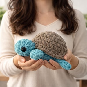 May include: A handmade crochet turtle plushie. The turtle has a brown shell and blue head, flippers, and tail. It has a black eye. The plushie is held in a person's hands.