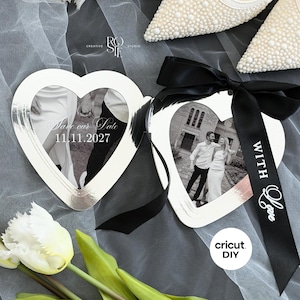 May include: Two heart-shaped silver frames featuring black and white photographs of a couple. One frame displays the text "Save our Date 11.11.2027" and the other is adorned with a black ribbon printed with the words "WITH Love".