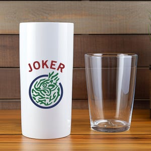 K&ouml;nnte beinhalten: Ein wei&szlig;er Becher mit dem Wort "JOKER" in roter Schrift und einer gr&uuml;n-blauen Grafik. Ein klares Glas befindet sich neben dem Becher. Beide stehen auf einer Holzoberfl&auml;che.