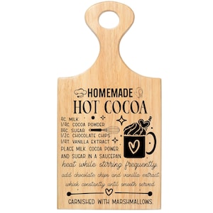 May include: Wooden cutting board with a handle, featuring a recipe for homemade hot cocoa. The recipe includes ingredients like milk, cocoa powder, sugar, chocolate chips, and vanilla extract. The board is decorated with illustrations and text.