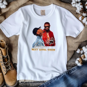 Pu&ograve; includere: Una t-shirt bianca a maniche corte con una stampa grafica di due uomini, uno con giacca di jeans e berretto nero, l'altro con camicia rossa e occhiali da sole. Il design include due firme bianche e il testo dorato NEXT LEVEL VISION.