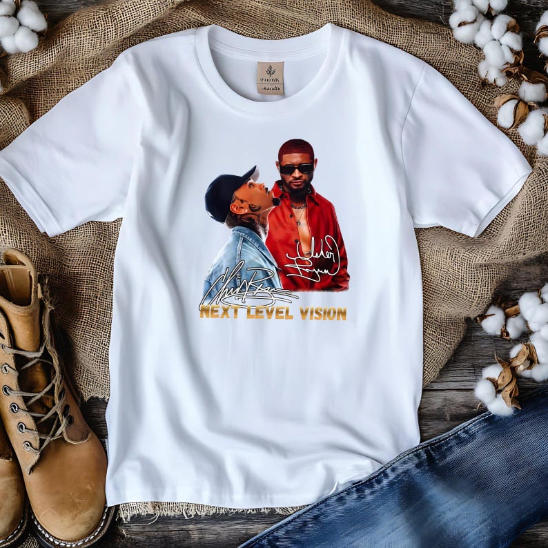 Pu&ograve; includere: Una t-shirt bianca a maniche corte con una stampa grafica di due uomini, uno con giacca di jeans e berretto nero, l'altro con camicia rossa e occhiali da sole. Il design include due firme bianche e il testo dorato NEXT LEVEL VISION.